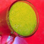 A glass mug filled with a green smoothie sits on a bright red surface.