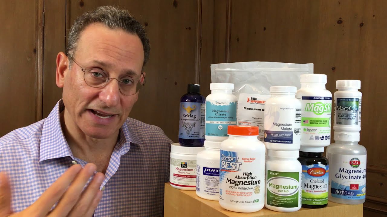 A person stands next to a variety of magnesium supplement bottles and containers displayed on a wooden surface.