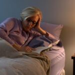 A person wearing glasses reads a book in bed next to a lit lamp and a glass of wine on the nightstand.