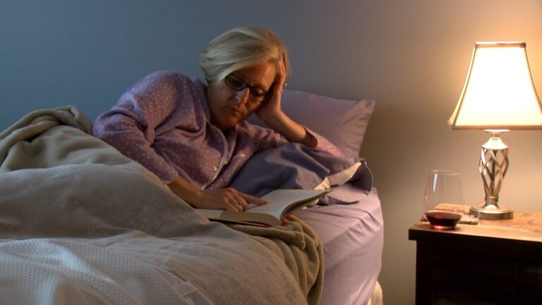 A person wearing glasses reads a book in bed next to a lit lamp and a glass of wine on the nightstand.